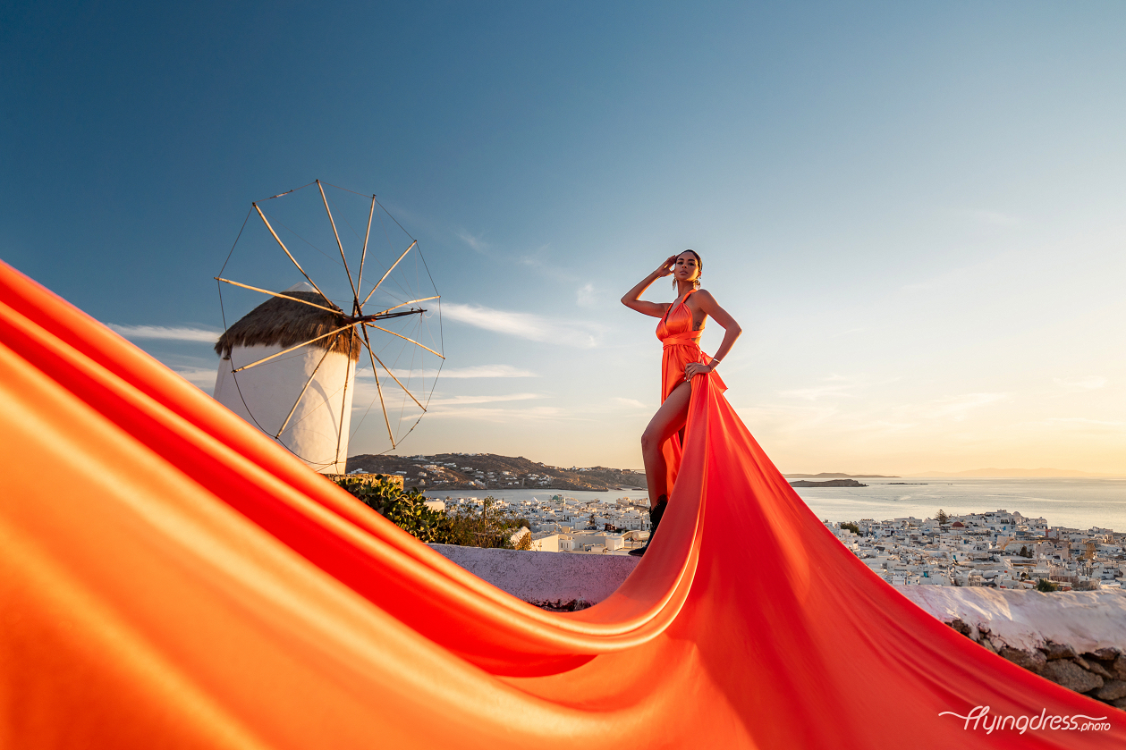 A woman in a flowing orange dress poses near Boni's Windmill in Mykonos, with the sunset casting a warm glow over the landscape and her dress trailing elegantly in the wind.